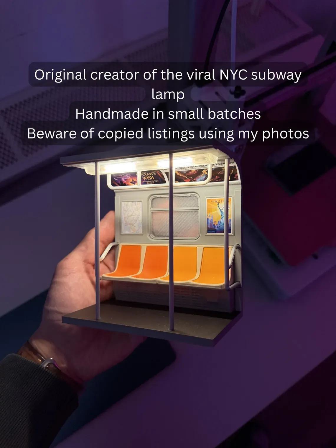 May include: A miniature NYC subway lamp with orange seats and posters. The lamp is handmade and features the text "Original creator of the viral NYC subway lamp". The lamp is held in a person